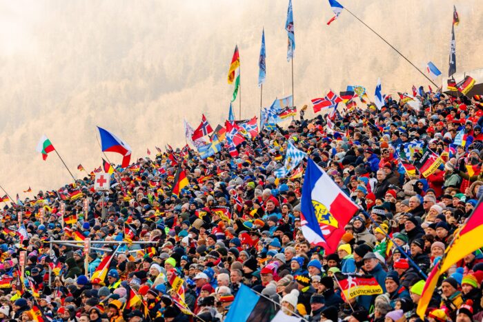 Audiodeskription-Biathlon-fuer-alle-erlebbar_gt2a6984_0 Großes Zuschauerfeld mit zahlreichen internationalen Fahnen feuert beim Biathlon in winterlicher Bergkulisse an.