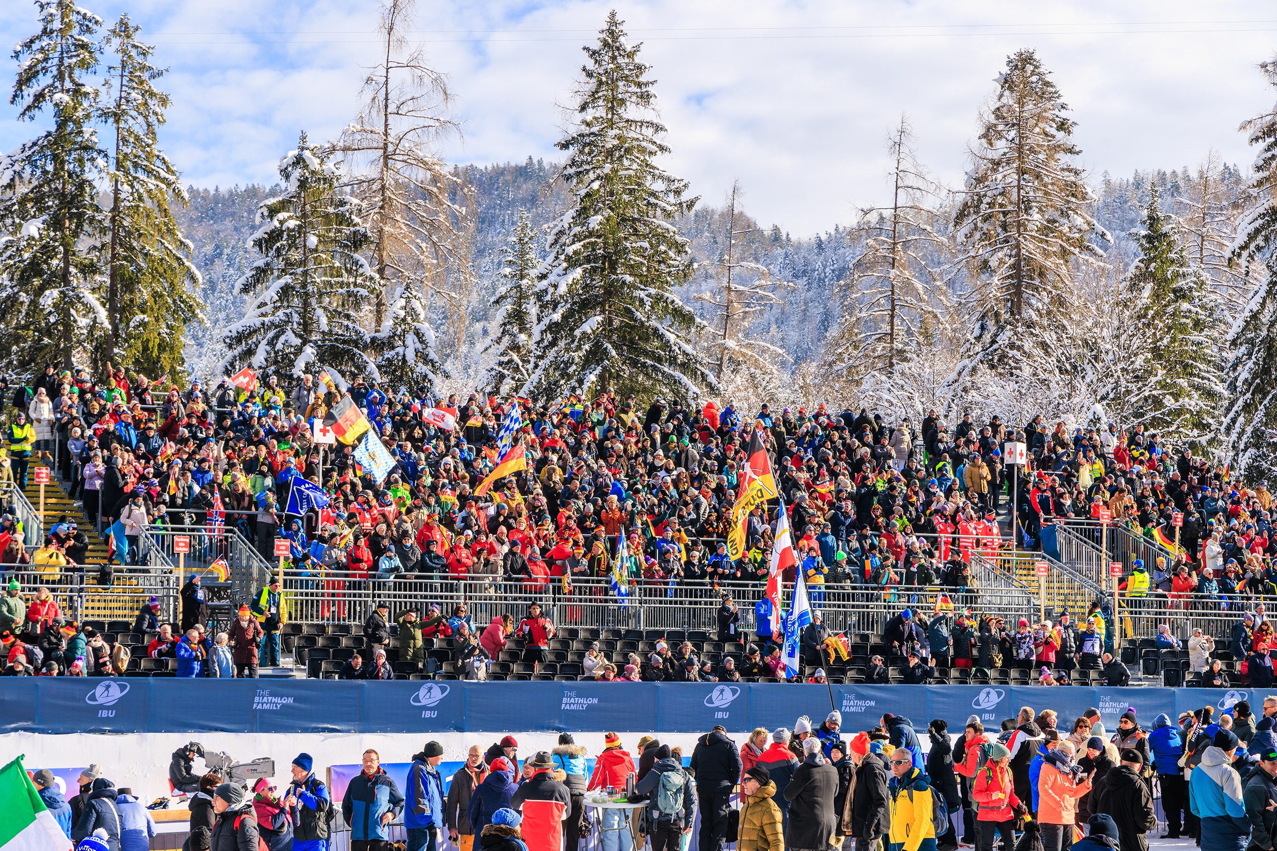 Große Zuschauertribüne mit Fans und Fahnen vor verschneiter Waldkulisse.