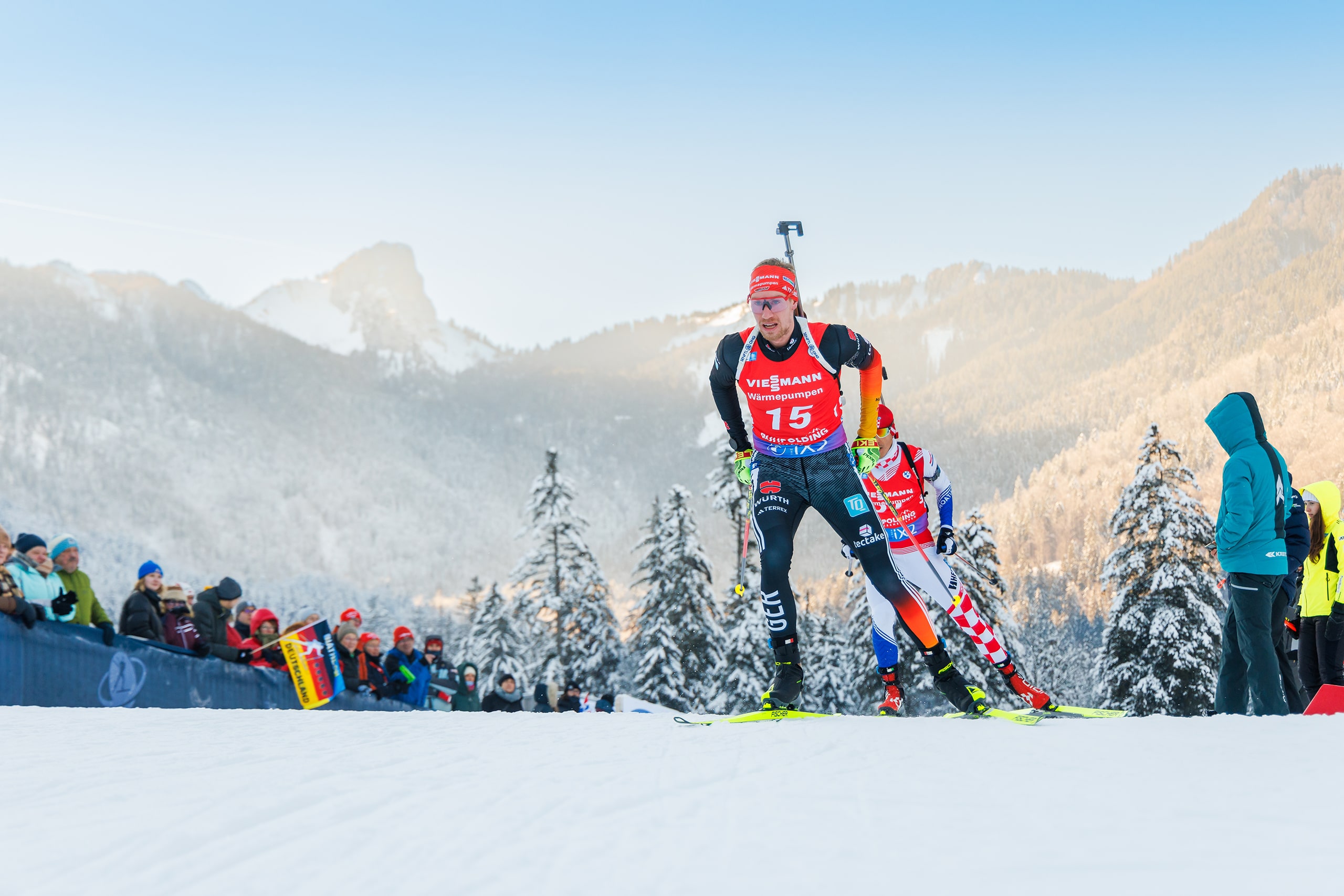 Biathlet mit Startnummer 15 läuft im Sprint vor winterlicher Bergkulisse, angefeuert von Zuschauern.