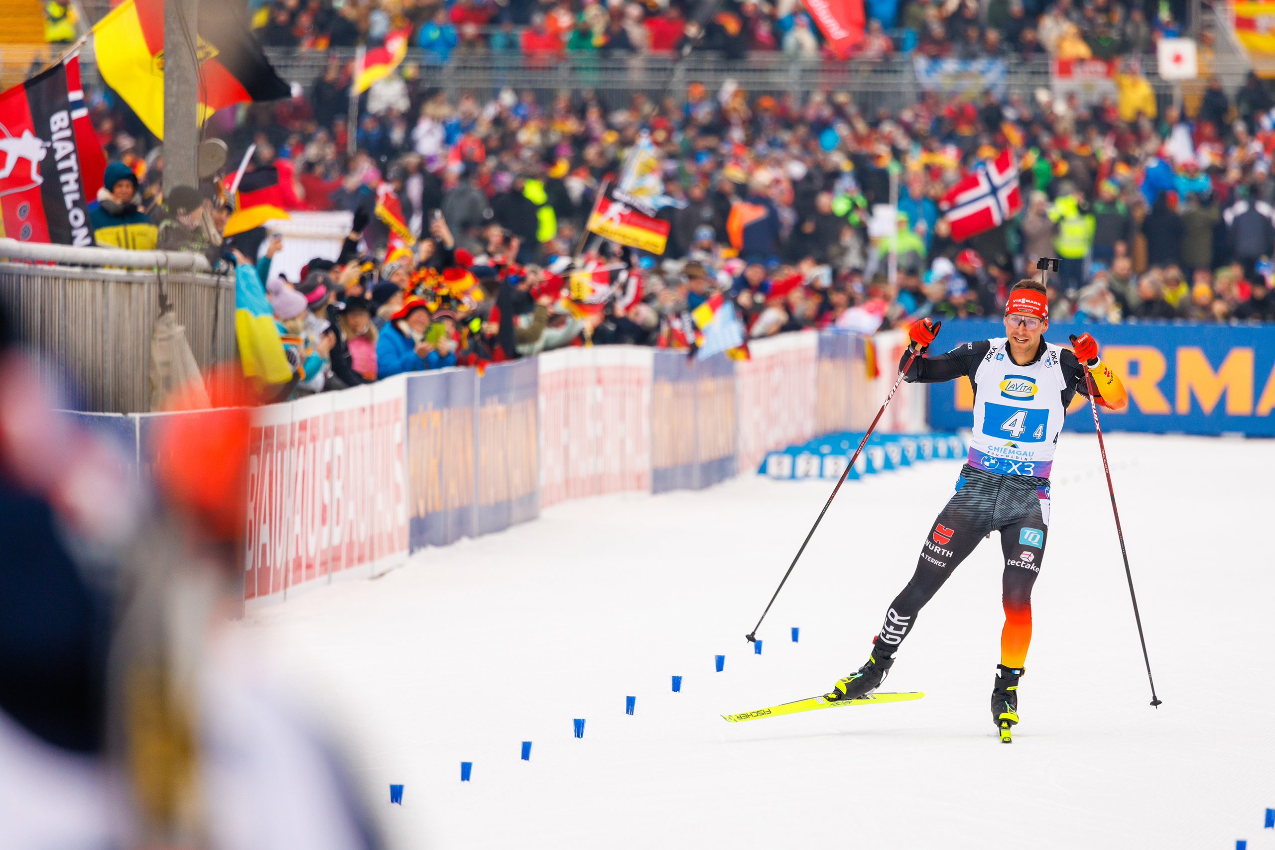 Biathlet mit Startnummer 44 läuft an jubelnden Fans im Stadionbereich der Chiemgau Arena vorbei.
