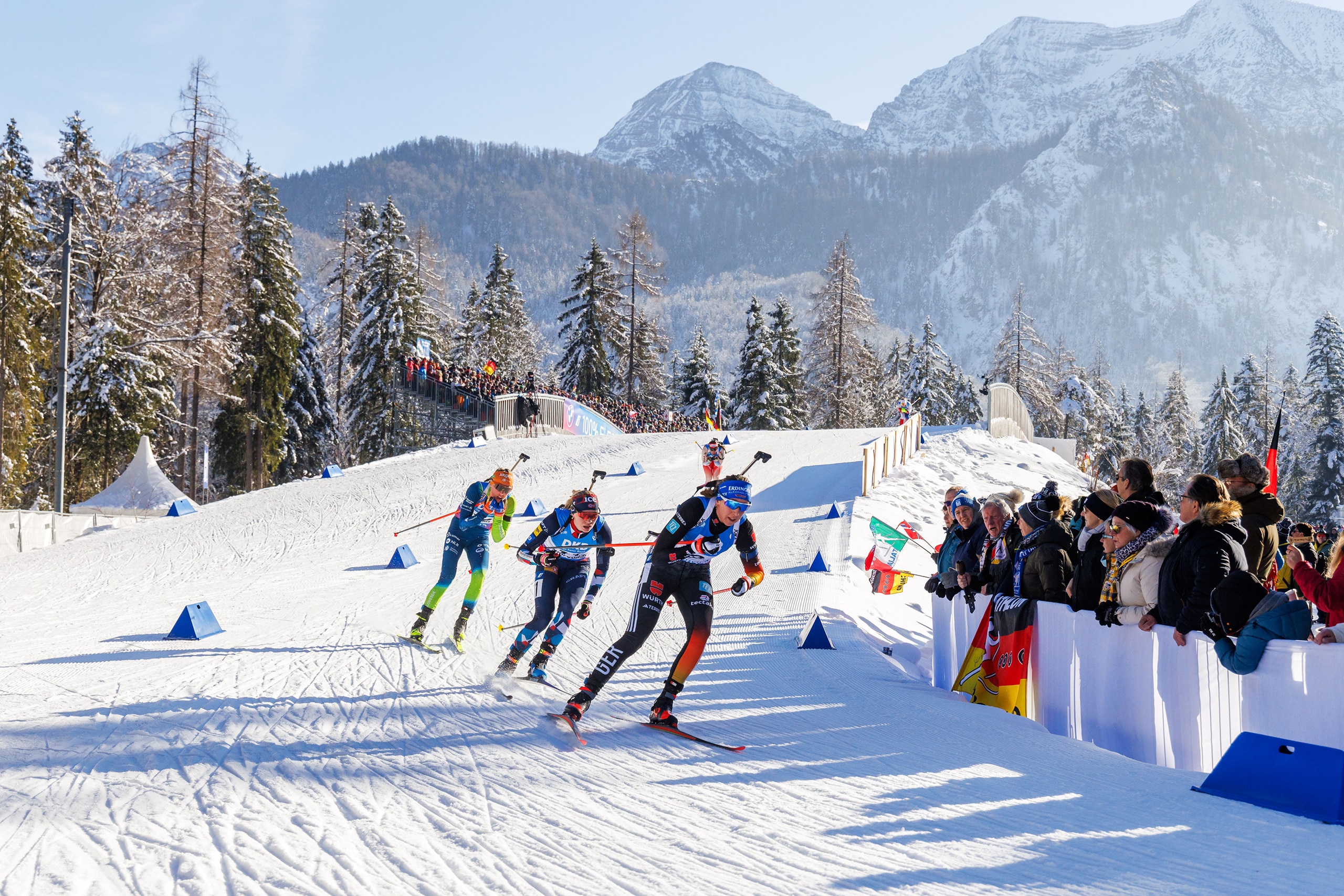 Mehrere Biathletinnen laufen in der Verfolgung eine Kurve hinunter, angefeuert von Zuschauern vor winterlicher Bergkulisse.