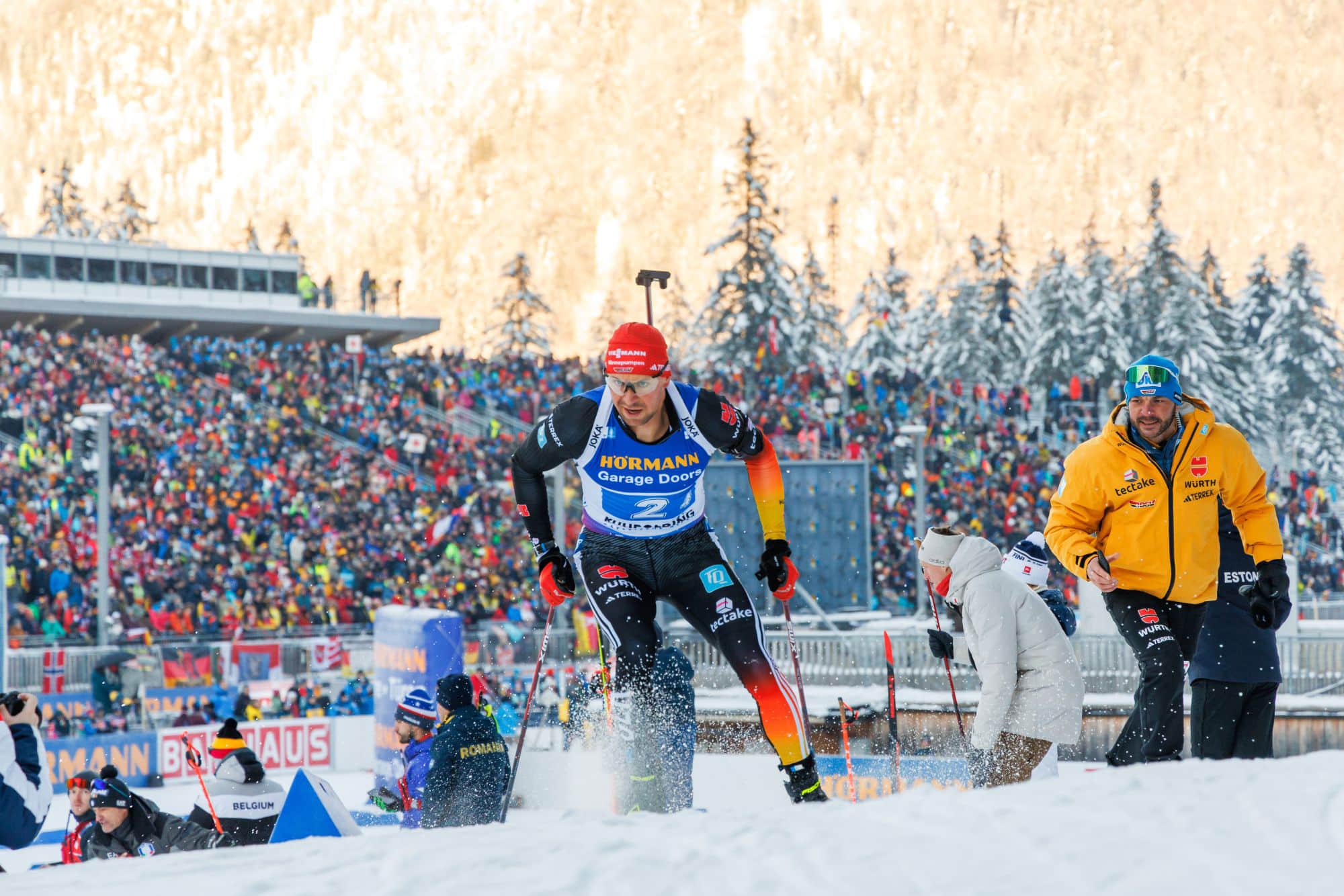 Biathlet in der Verfolgung kämpft sich eine Steigung hinauf vor einer großen Zuschauertribüne.