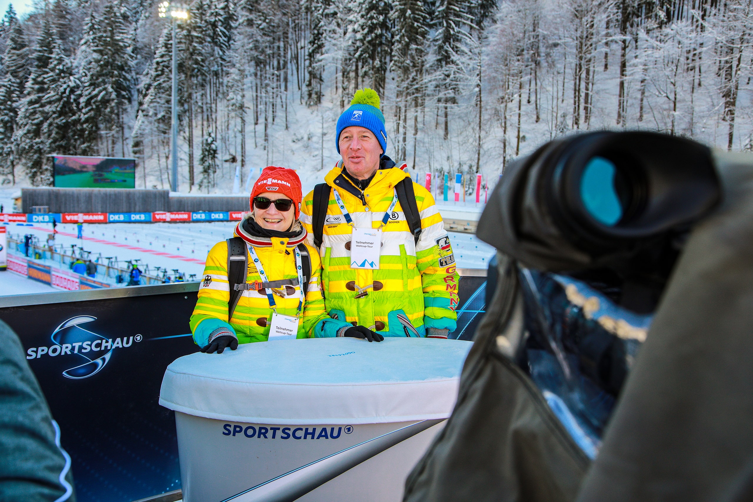 Zwei Zuschauer in gelben Winterjacken stehen im Stadionbereich der Chiemgau Arena vor einer Sportschau-Plattform.