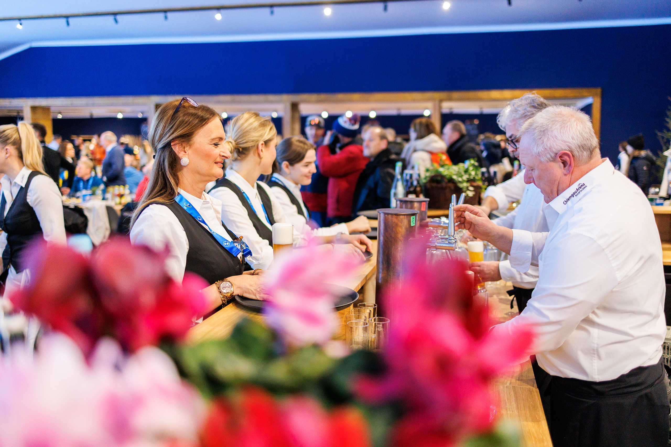 VIP_GT2A8503 Servicekräfte im VIP-Bereich schenken an der Bar Getränke aus, im Hintergrund sitzen und stehen Gäste an den Tischen.