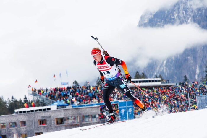 Verfolgung_Maenner_GT2A3497 Biathlet mit Startnummer 13 läuft in der Verfolgung vor vollbesetzten Tribünen und Bergkulisse.