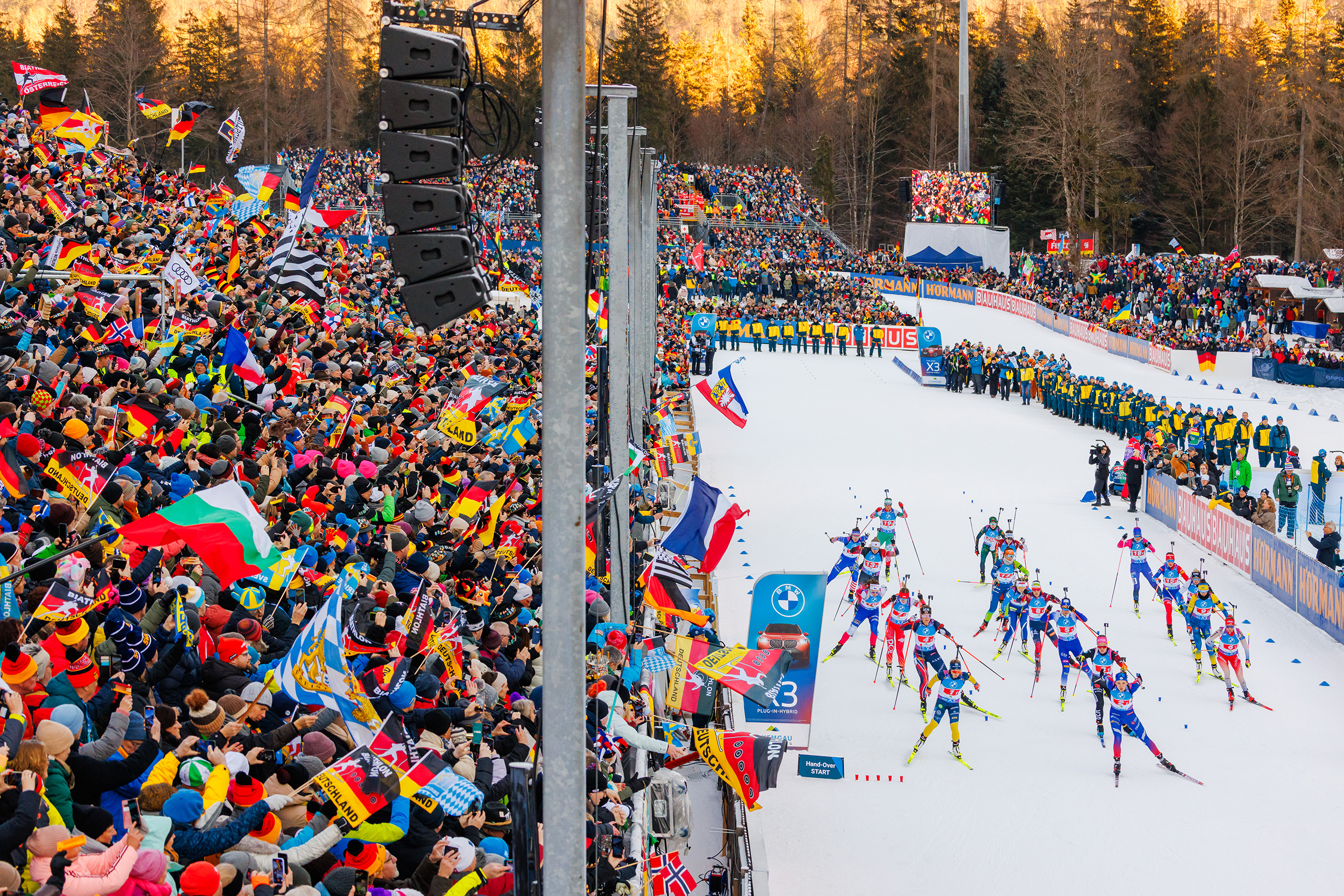 Weltcup-2027_Header_GT2A8808 Startfeld der Biathlonstaffel mit zahlreichen Athleten, dicht gefüllten Zuschauertribünen und vielen Fahnen in der Chiemgau Arena.
