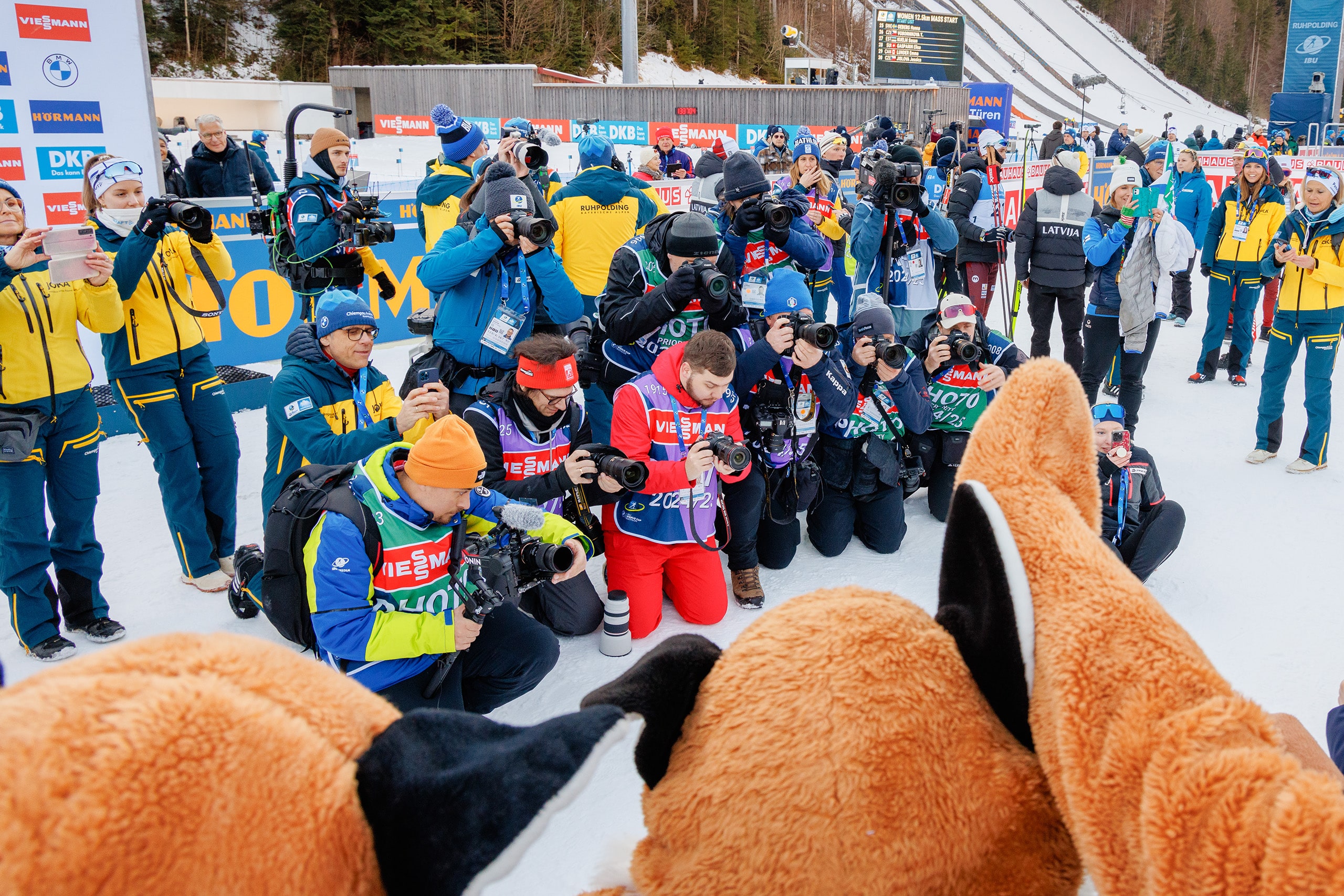 Pressefotografen knien im Zielbereich der Chiemgau Arena und fotografieren Maskottchen beim Biathlon-Weltcup in Ruhpolding.
