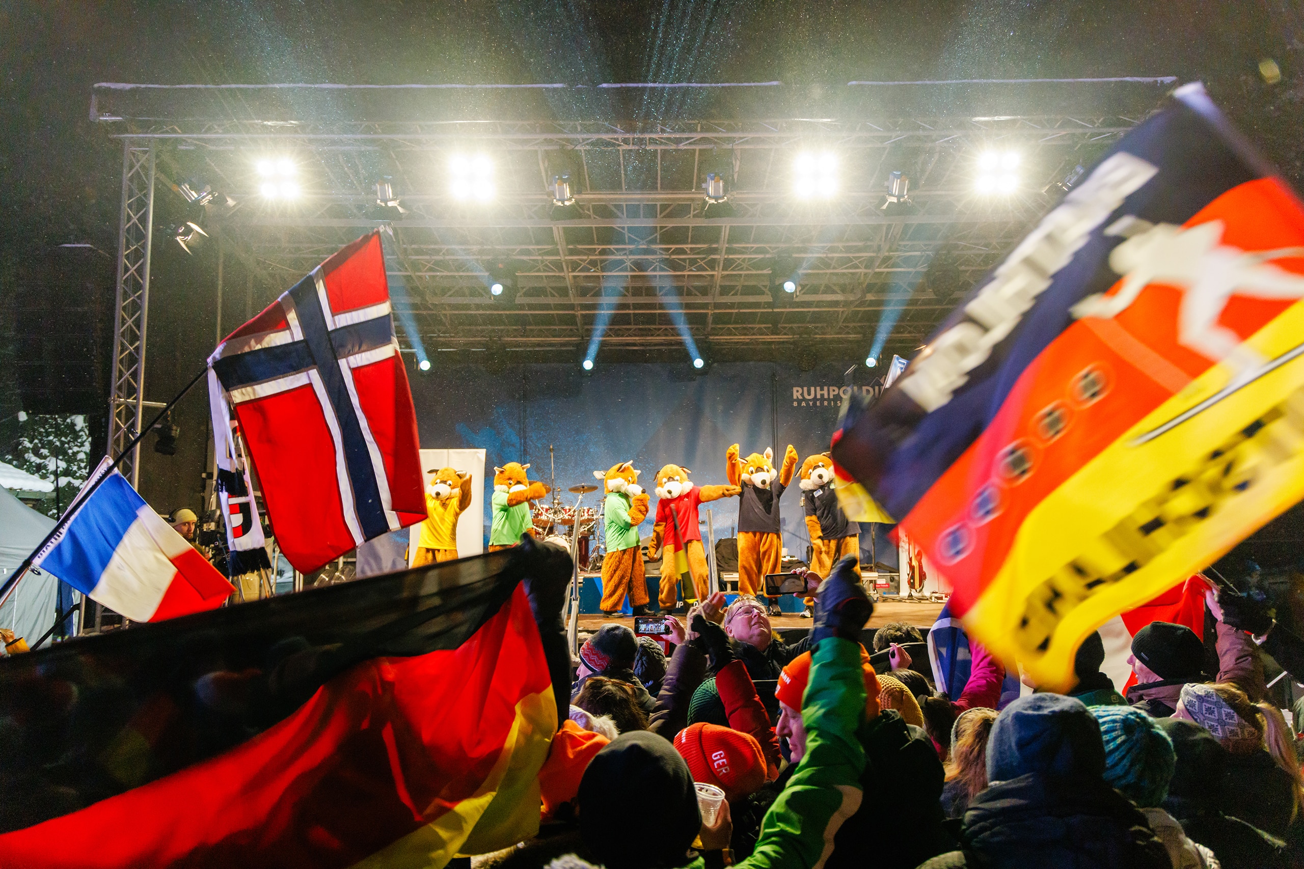Maskottchen tanzen auf der Bühne, während die Menge mit internationalen Fahnen jubelt.