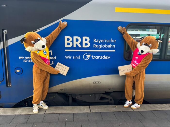 brb_beppo_30 Zwei Beppo-Fuchs-Maskottchen stehen vor einem BRB-Zug und halten Schilder zum Biathlon-Weltcup 2026 in Ruhpolding hoch.