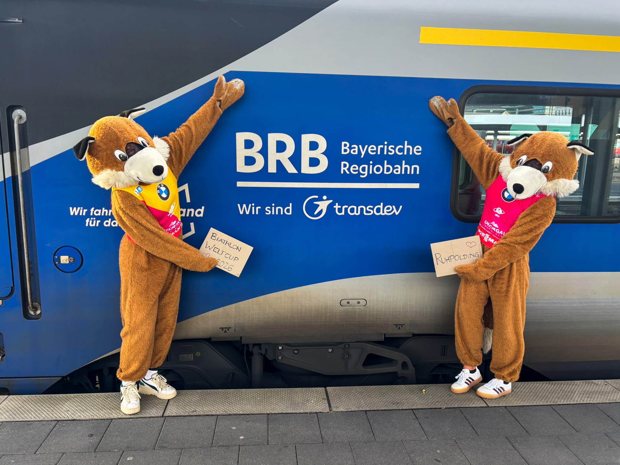 Zwei Beppo-Fuchs-Maskottchen stehen vor einem BRB-Zug und halten Schilder zum Biathlon-Weltcup 2026 in Ruhpolding hoch.